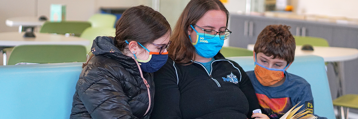 Mom and two kids with masks on reading a book in the library